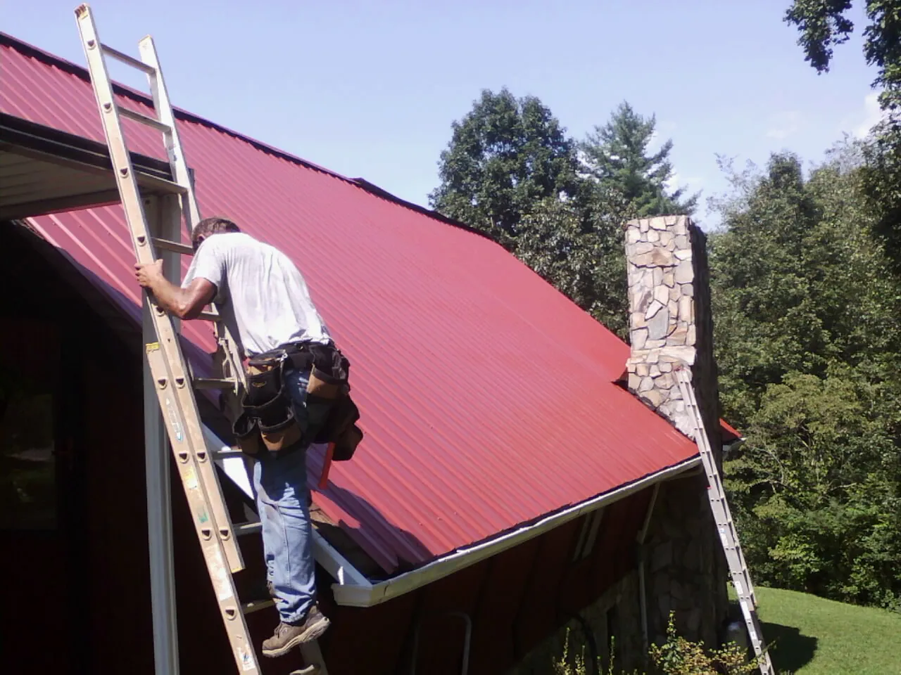 Roofer on a ladder beside a red metal roof and stone chimney — Soffit & Fascia Repair by Roofing Brothers in Perrysburg